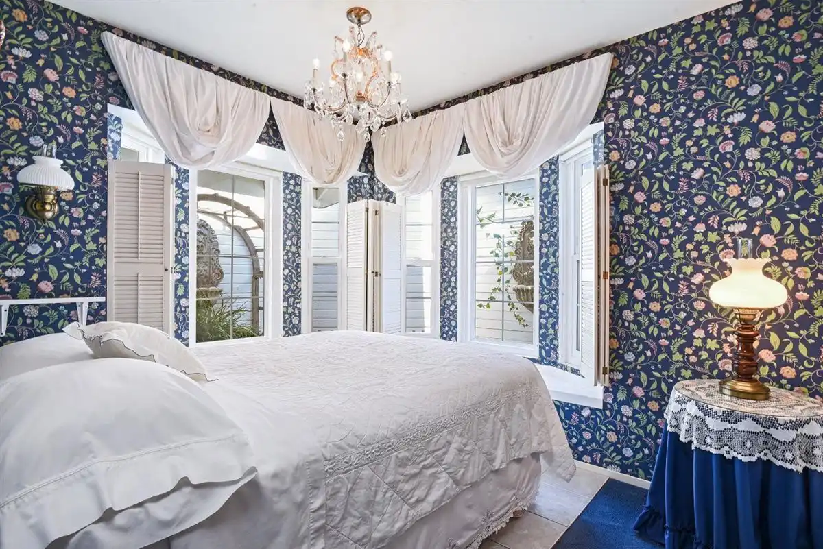 A crystal chandelier hangs over windows with white balloon valance curtains, full bed, and dark blue floral wallpaper.